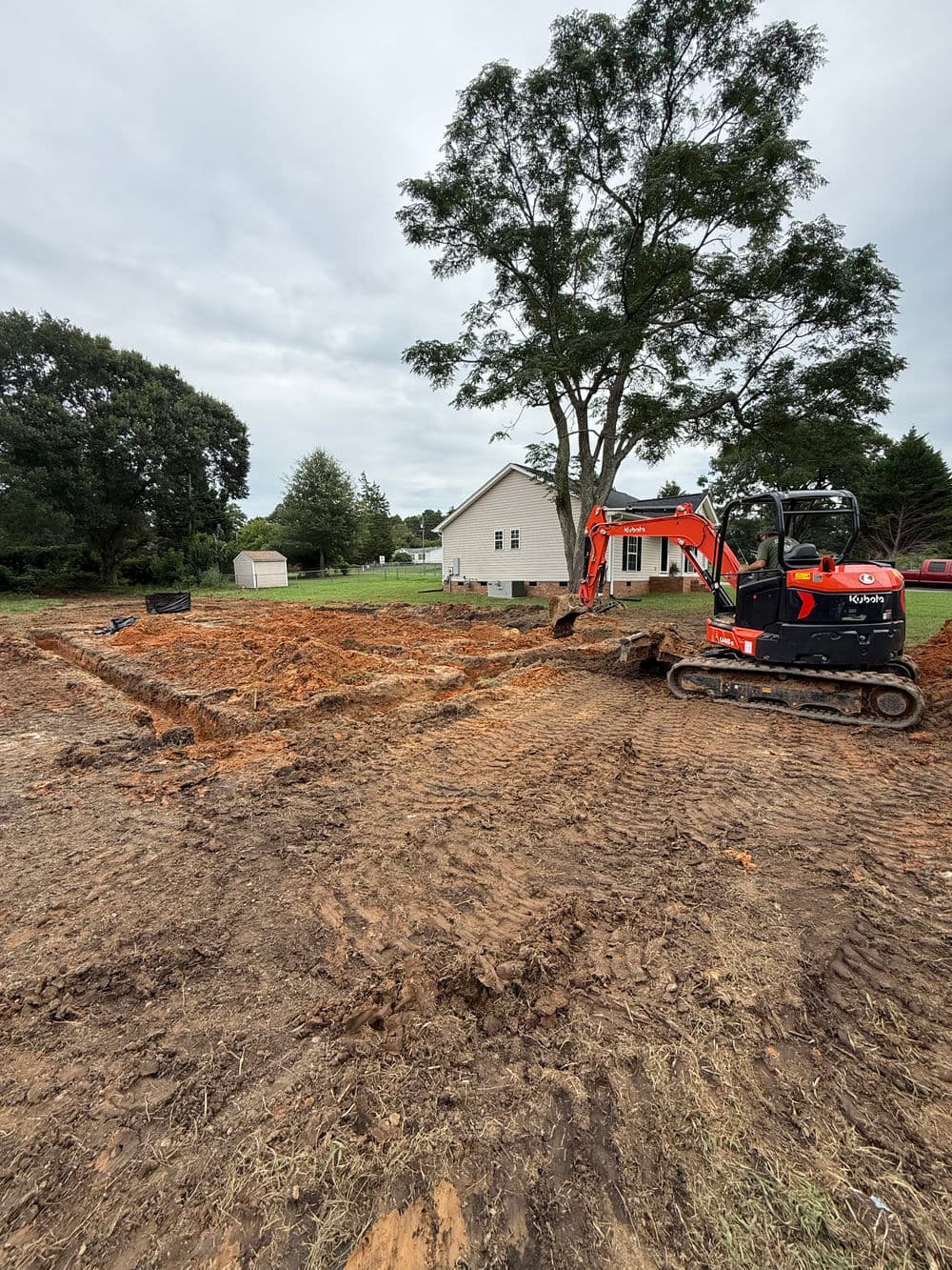 Excavator working on a construction site with a house and tree in the background.