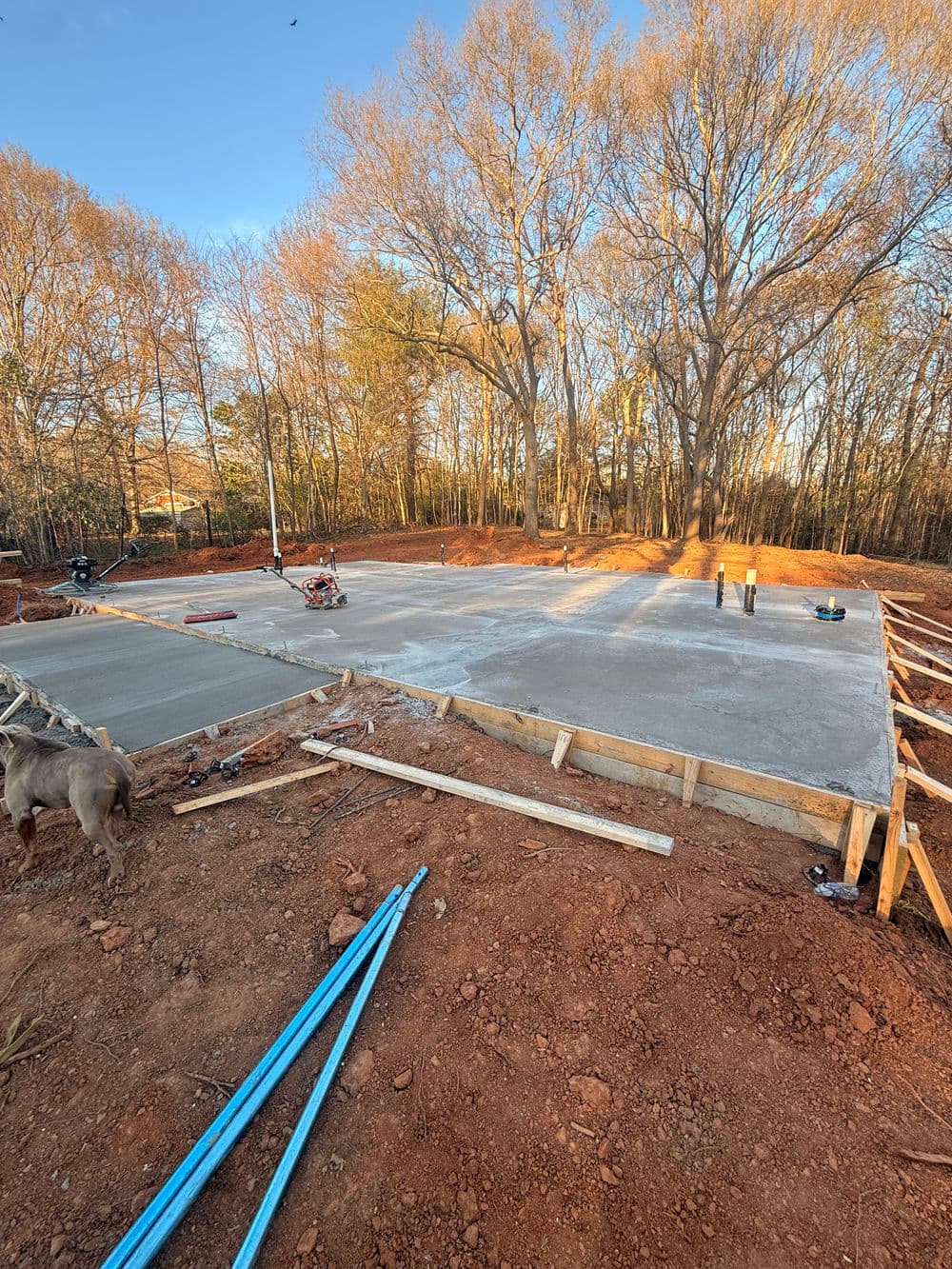 Newly poured concrete foundation surrounded by trees, with construction tools and equipment.