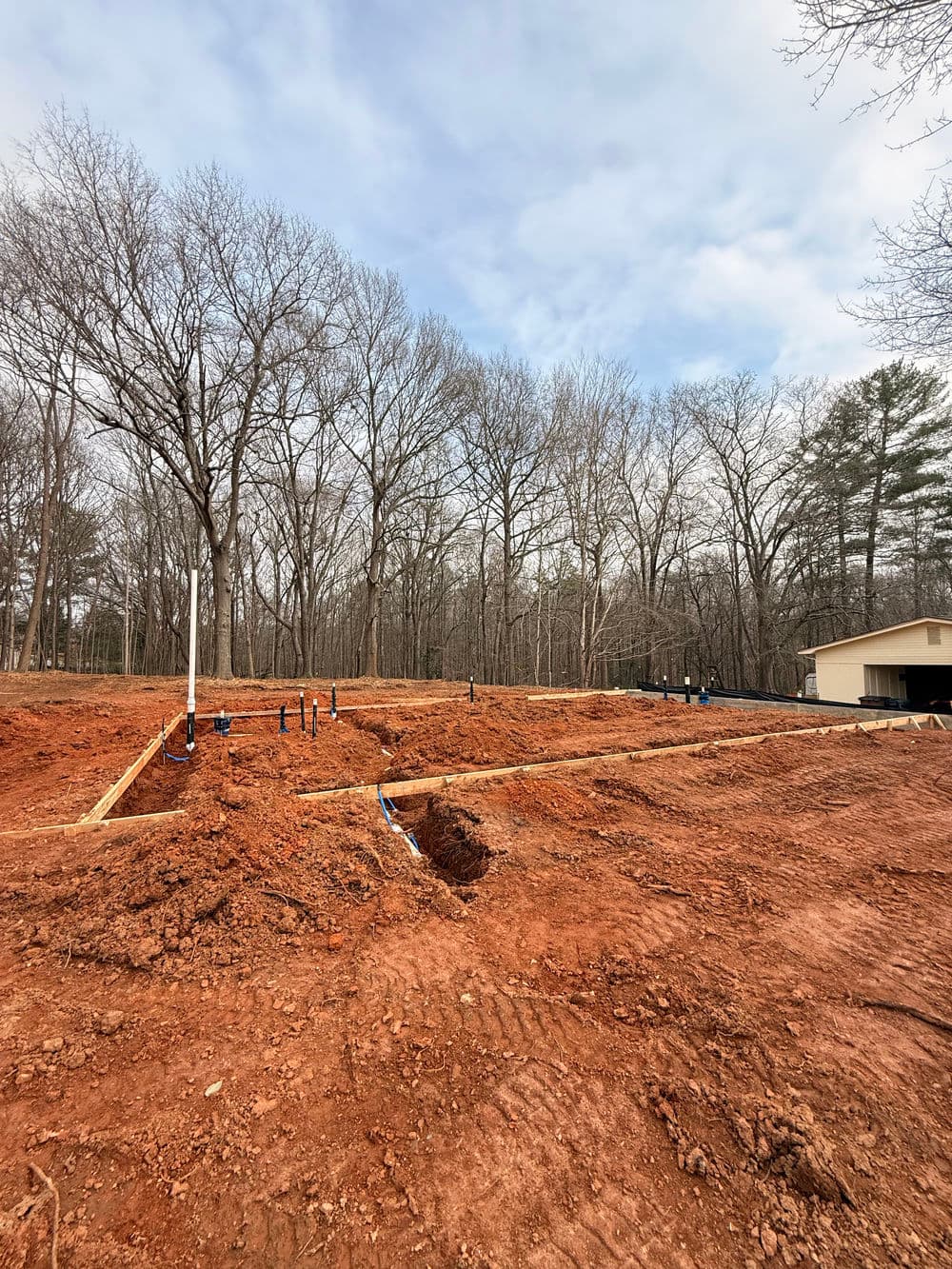 Construction site with red dirt, plumbing pipes, and wooden framing in a wooded area.