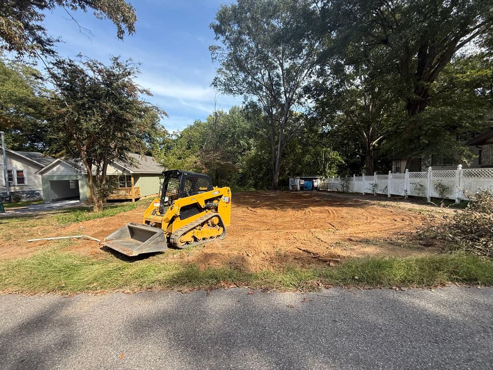 Yellow compact track loader on a cleared lot with trees and fence in the background.