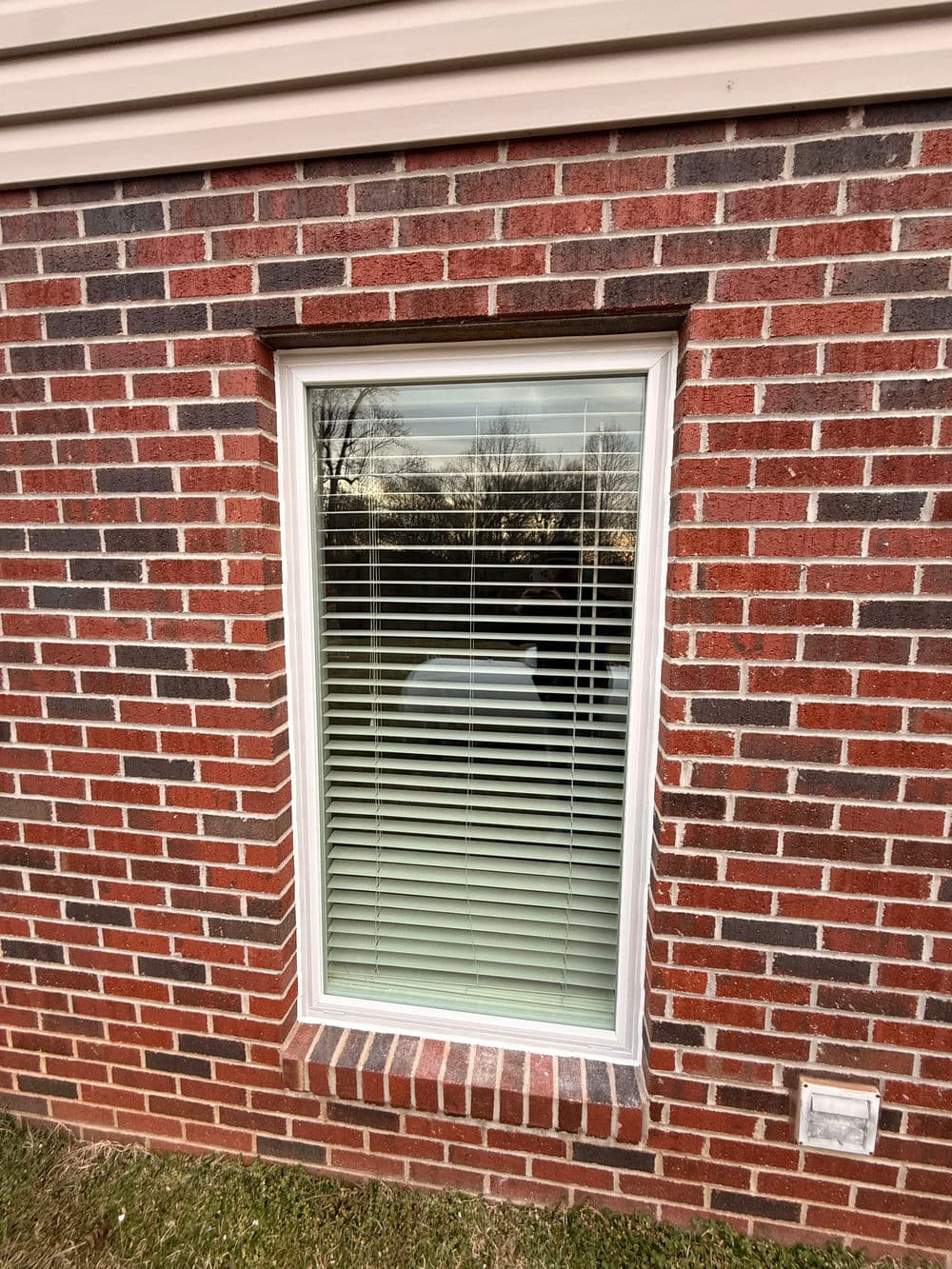 Rectangular window with white blinds set in a brick wall, reflecting outdoor scenery.