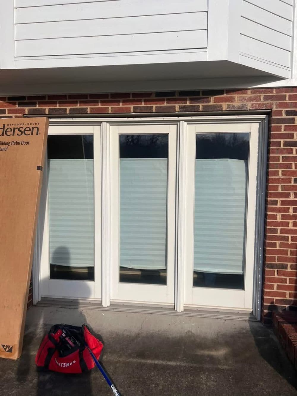 Andersen sliding patio door installation with weather protection and tools on a brick exterior.