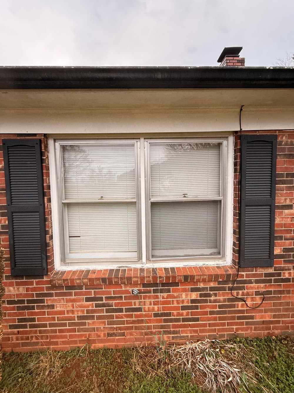 Brick house exterior with two windows, black shutters, and grassy area below.