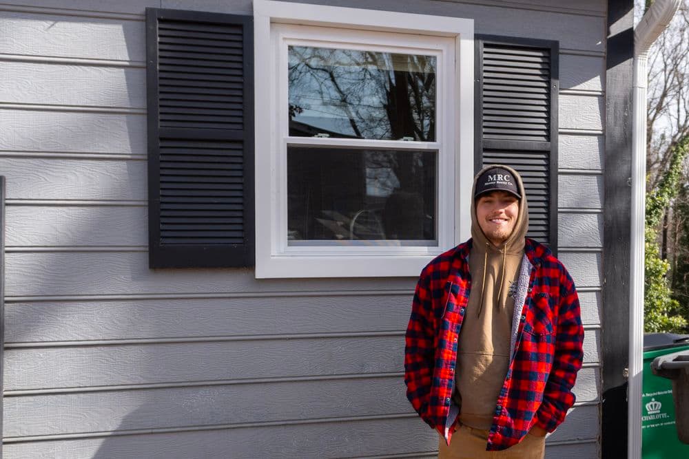 Young man in plaid jacket standing by a gray house with a window and shuttered exterior.