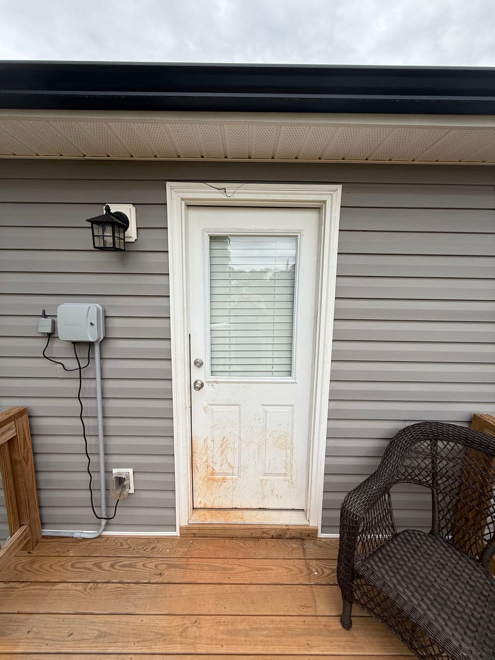 Front door with dirty glass, gray siding, light fixture, and wooden deck furniture.