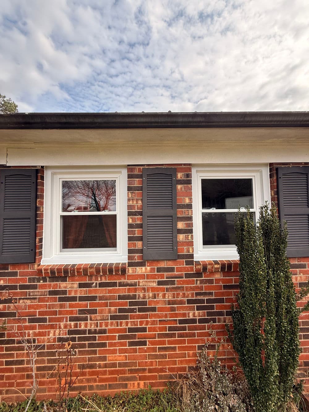 Brick house exterior with two windows and shutters under a cloudy sky.