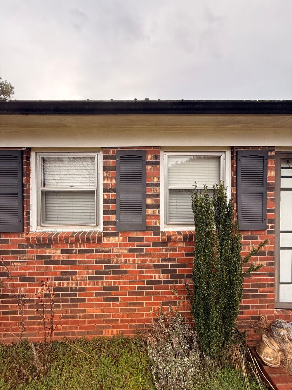 Brick house exterior with two windows and shutters, featuring greenery in front.