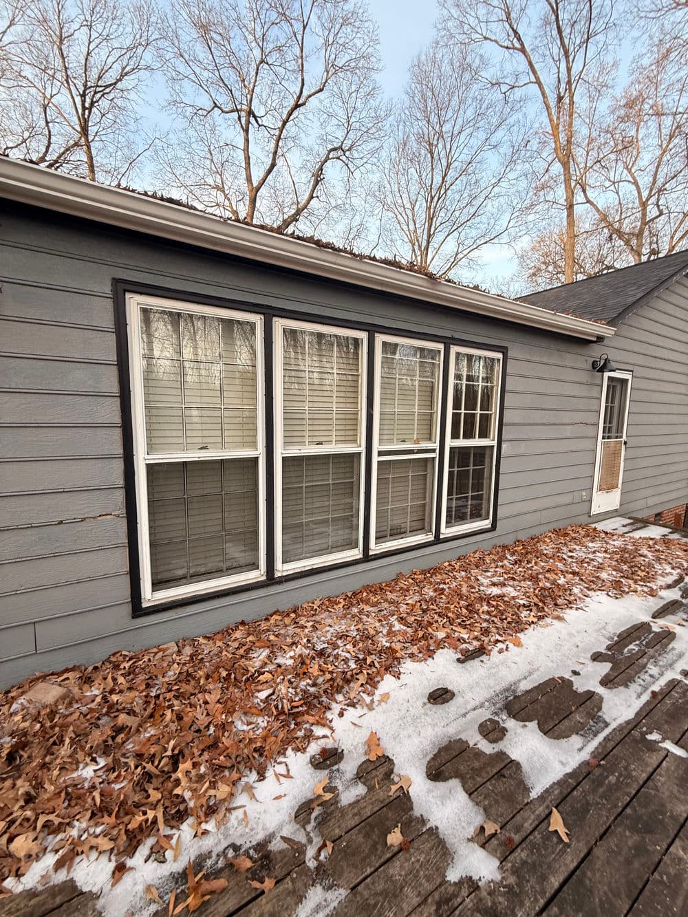 Gray house exterior featuring large windows, fallen leaves, and a snowy deck.